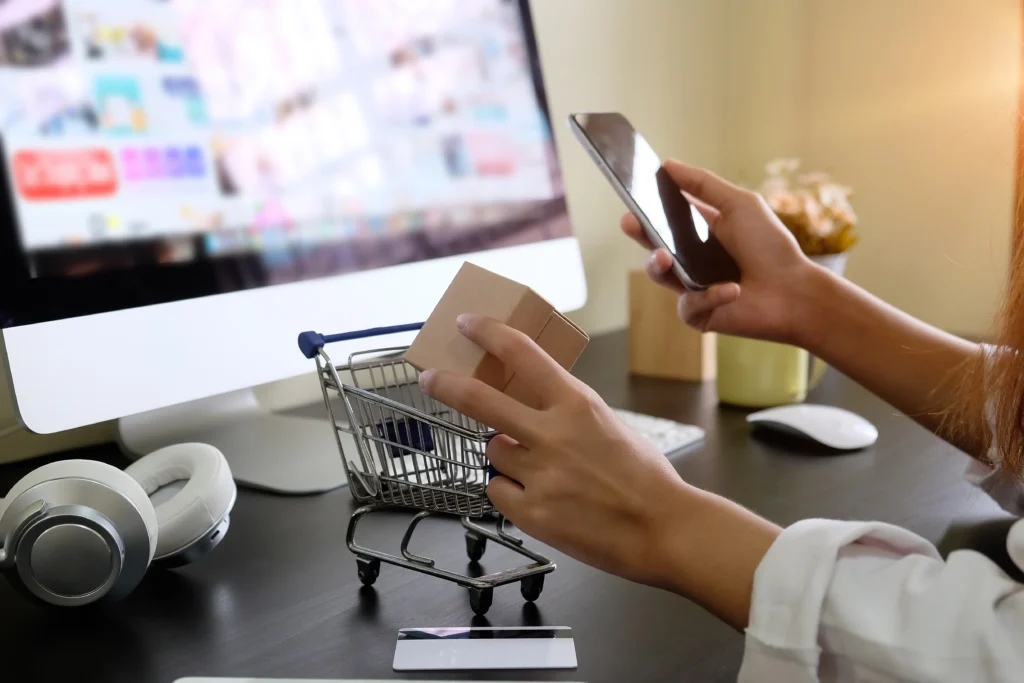 Mujer con un celular en la mano y una caja pequeña frente a un computador de escritorio haciendo una compra por internet